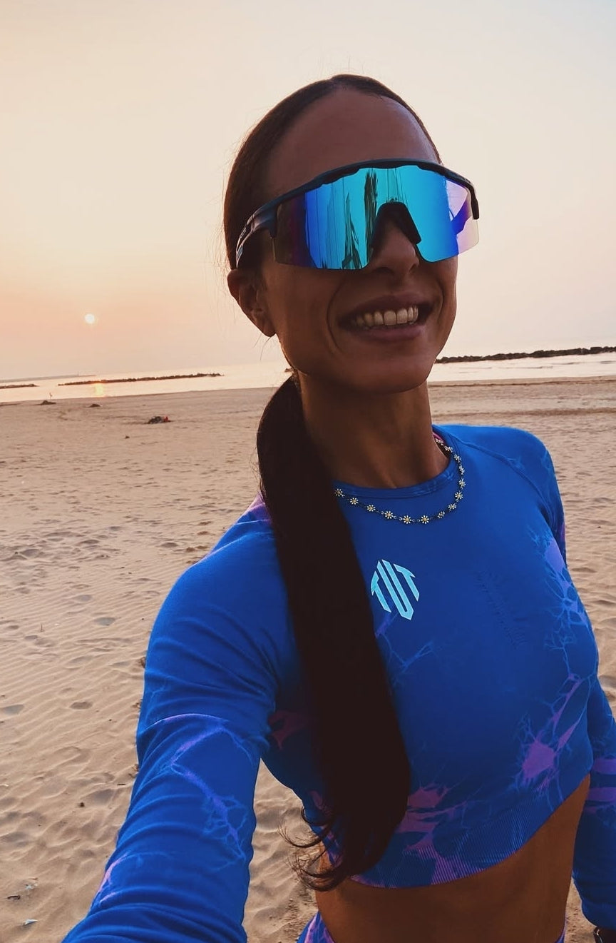 Person wearing blue sunglasses and a blue shirt on a beach at sunset