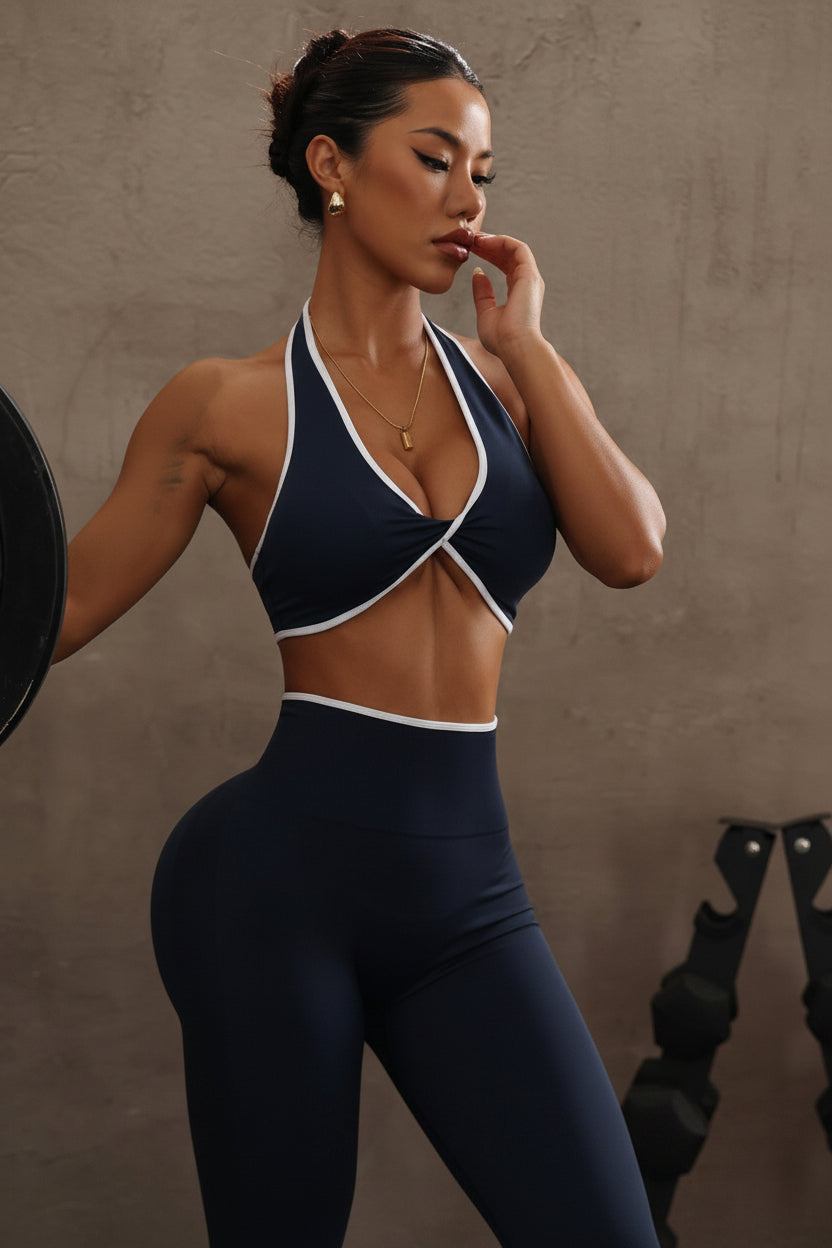 A woman posing in a gym setting wearing a navy blue sports bra and matching high-waisted leggings with white trim, holding a white smartphone to take a mirror selfie.