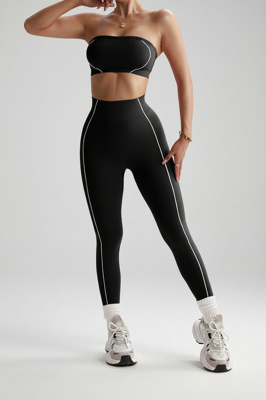 A woman modeling a matching black activewear set consisting of a strapless bandeau crop top and high-waisted leggings with thin white side piping, paired with white socks and white sneakers, against a plain white background.