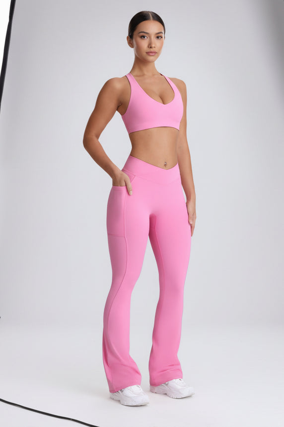 A woman modeling a bright pink activewear set consisting of a sports bra and high-waisted flared leggings with side pockets, paired with white sneakers, against a light gray studio background.
