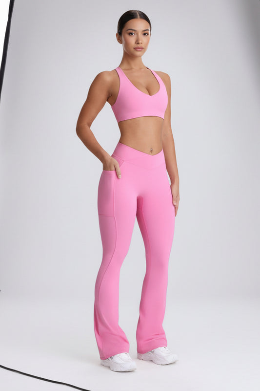 A woman modeling a bright pink activewear set consisting of a sports bra and high-waisted flared leggings with side pockets, paired with white sneakers, against a light gray studio background.