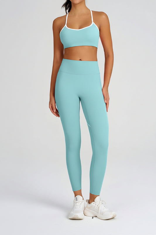 A woman modeling a matching light blue activewear set consisting of a sports bra with white trim and high-waisted leggings, paired with white sneakers, against a plain light gray studio background.