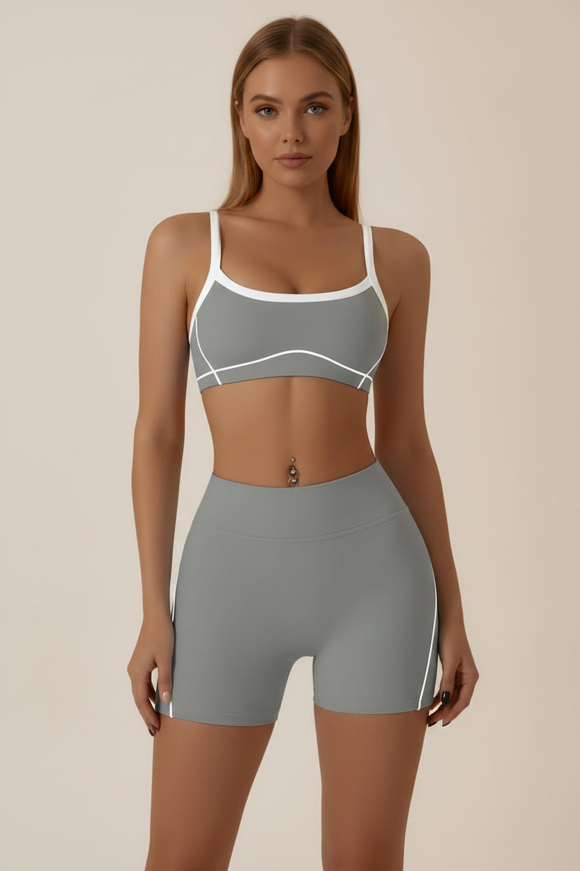 A woman modeling a gray athletic set consisting of a sports bra and matching high-waisted shorts with white contrast trim and side stripes, against a plain light gray studio background.