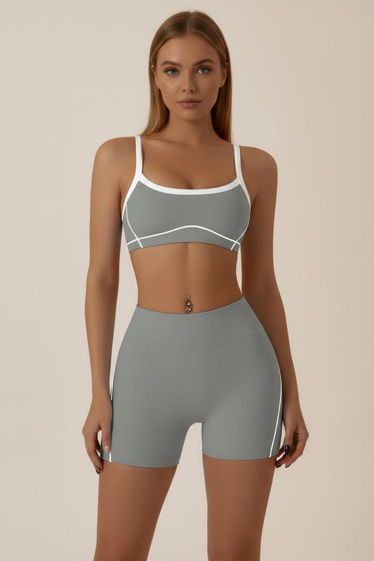 A woman modeling a gray athletic set consisting of a sports bra and matching high-waisted shorts with white contrast trim and side stripes, against a plain light gray studio background.