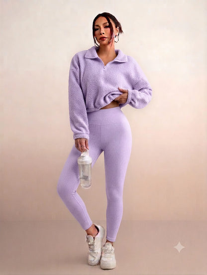A woman modeling a matching lavender fleece quarter-zip pullover and high-waisted leggings set, holding a clear water bottle, against a solid beige studio background.