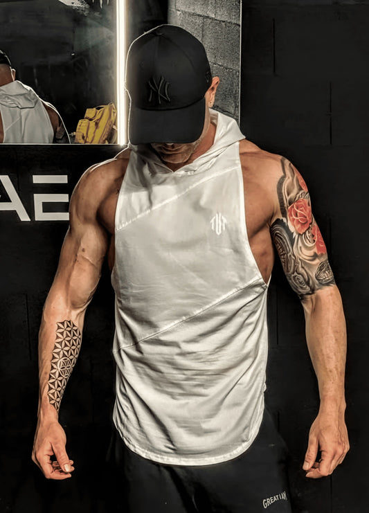 Man wearing a white sleeveless hoodie and black cap with tattoos on arms, standing against a dark background.