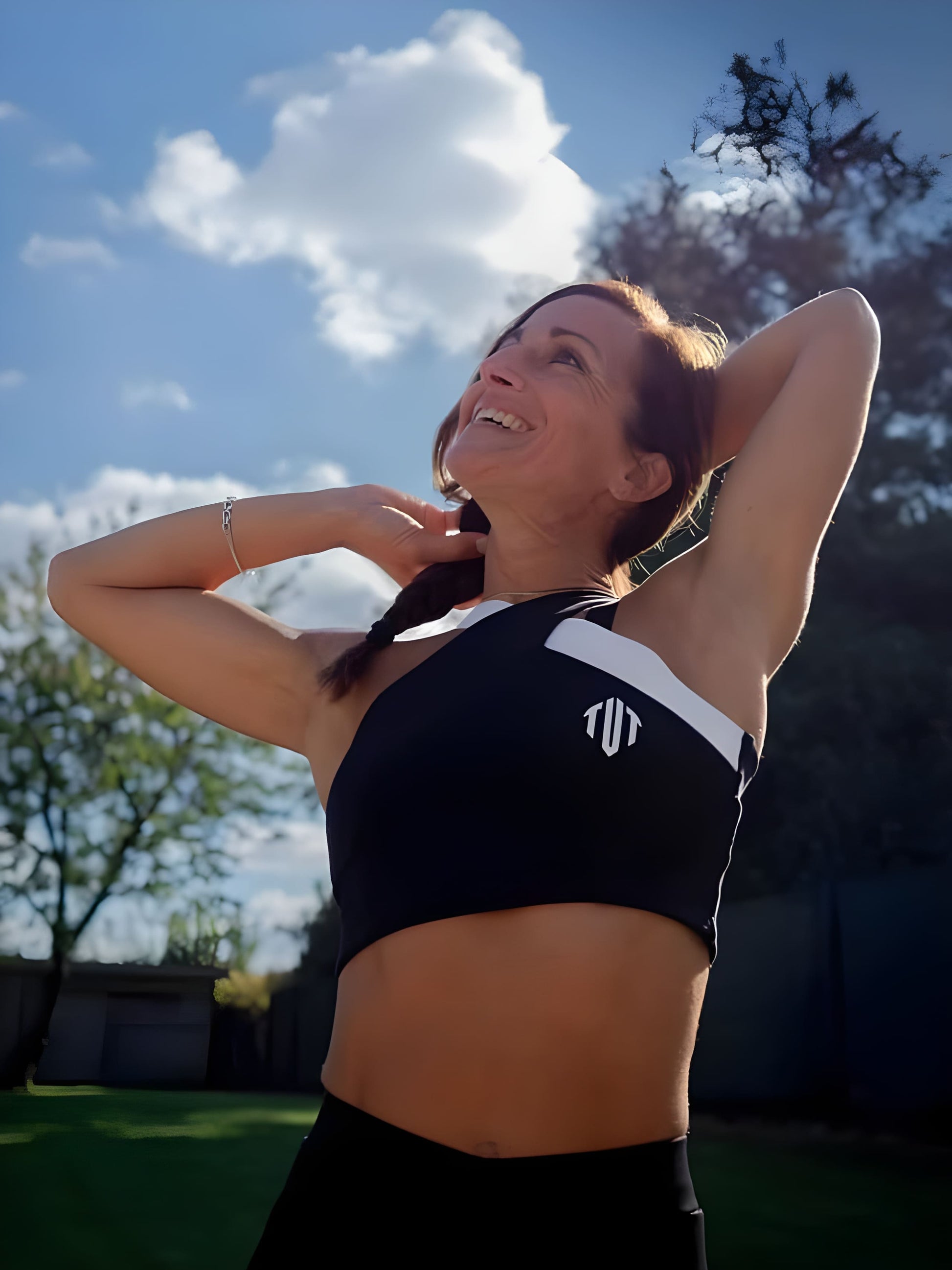 Woman in black sports bra and shorts stretching outdoors with trees and blue sky in the background